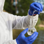 Water Purity Test. Hand holding a chemical flask or test tube with water. Lake or river in the background.